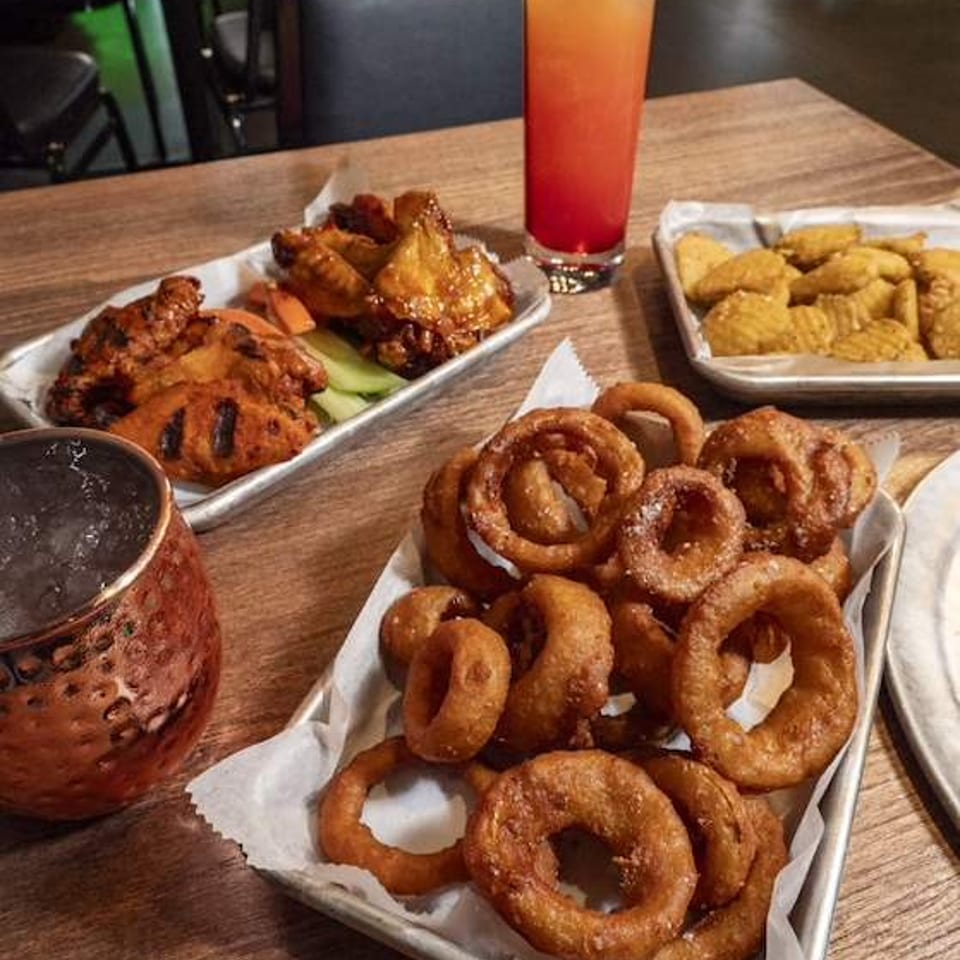 Wooden table laden with crispy onion rings, fried chicken, fries, dipping sauces, and a colorful cocktail with a parasol.