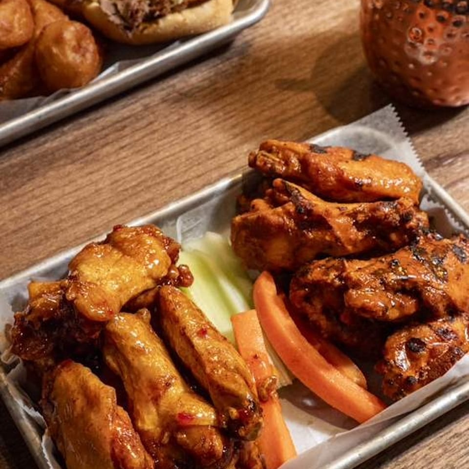 Metal tray of glazed chicken wings and grilled chicken with celery and carrot sticks, alongside a sandwich, fries, and copper cocktail.
