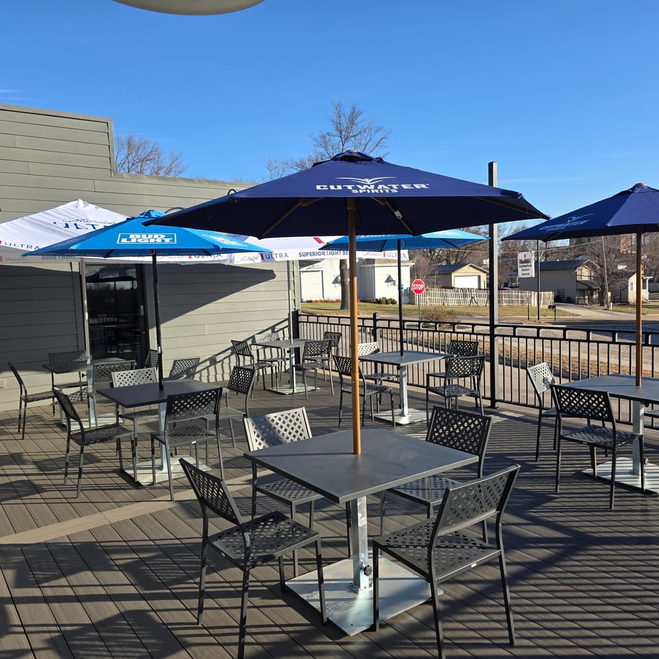 Outdoor restaurant patio with empty tables, lattice chairs, and branded umbrellas overlooking a sunny residential street.