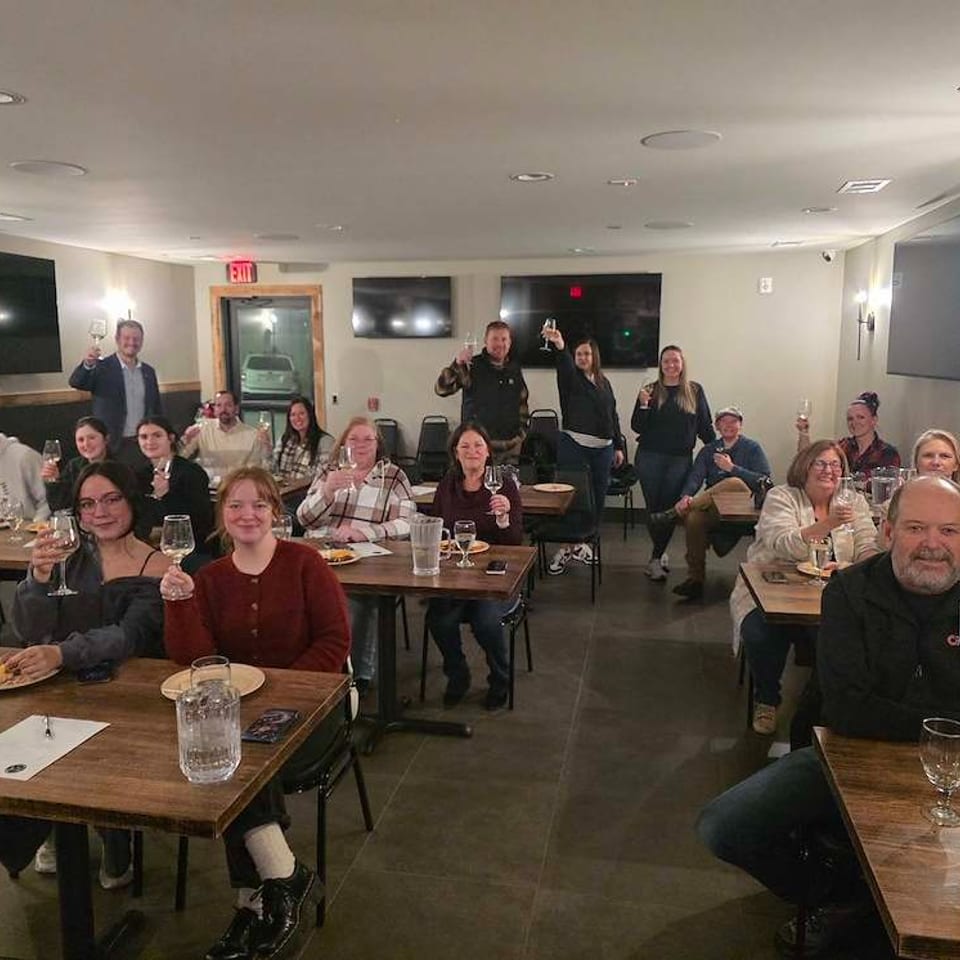 Group of diners raise wine glasses and smile inside a warmly lit restaurant dining room.