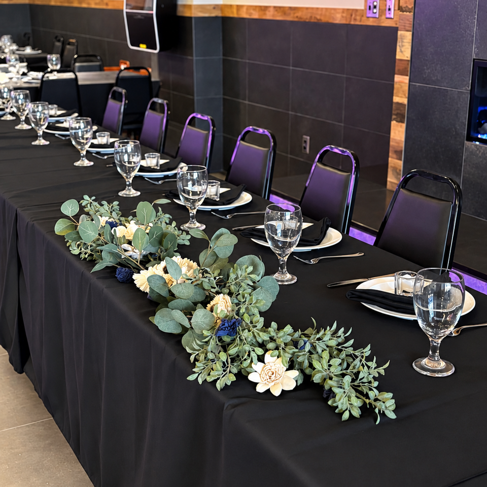 Long banquet table dressed in black linens, place settings, wine glasses, and a greenery-and-flower centerpiece in a purple-lit restaurant.