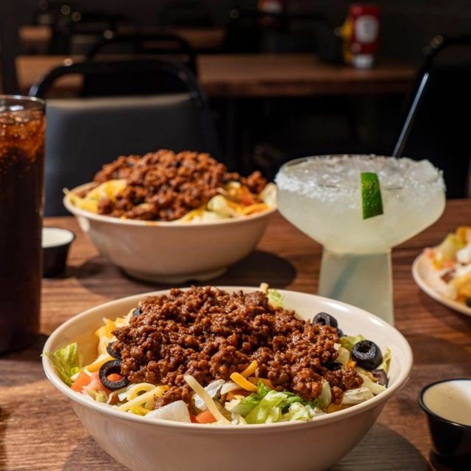 Taco salad bowls topped with seasoned ground beef sit on a wooden table beside a salted lime margarita and iced drink.