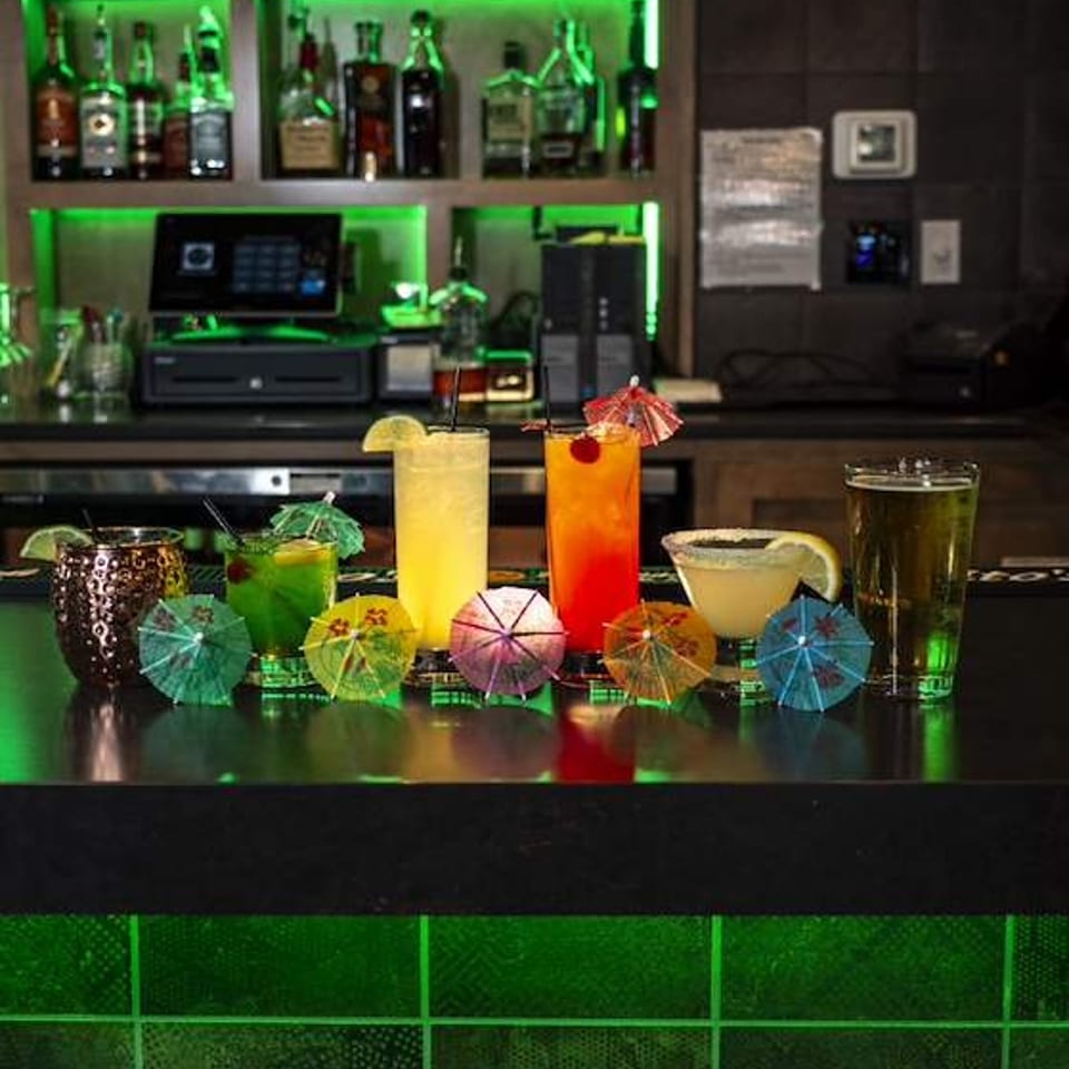 Colorful cocktails garnished with citrus, straws, and umbrellas line a bar beneath green-lit liquor shelves.
