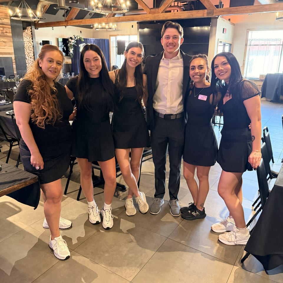 Six smiling restaurant staff members pose together inside a warmly lit dining room with exposed wooden beams and chandeliers.