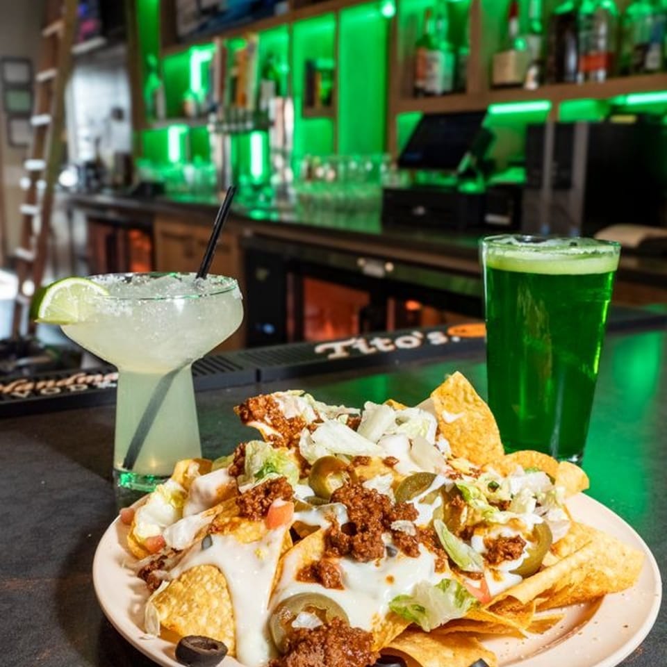 Loaded nachos with beef, cheese, jalapeños, and lettuce beside a margarita and green drink at a neon-lit bar.