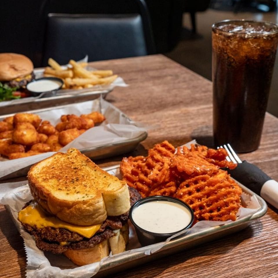 Restaurant table with a stacked cheeseburger, waffle fries, dipping sauce, fried bites, and a tall iced cola.