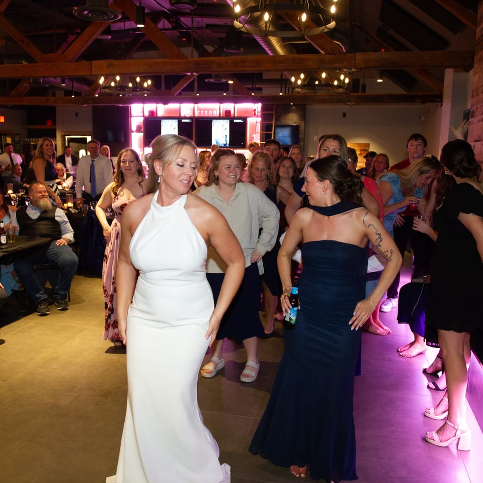 Guests dance and socialize inside a warmly lit restaurant with exposed beams, chandeliers, tables, and purple accent lighting.